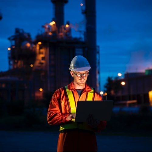Technician checking laptop outside at night with refinery structures in background